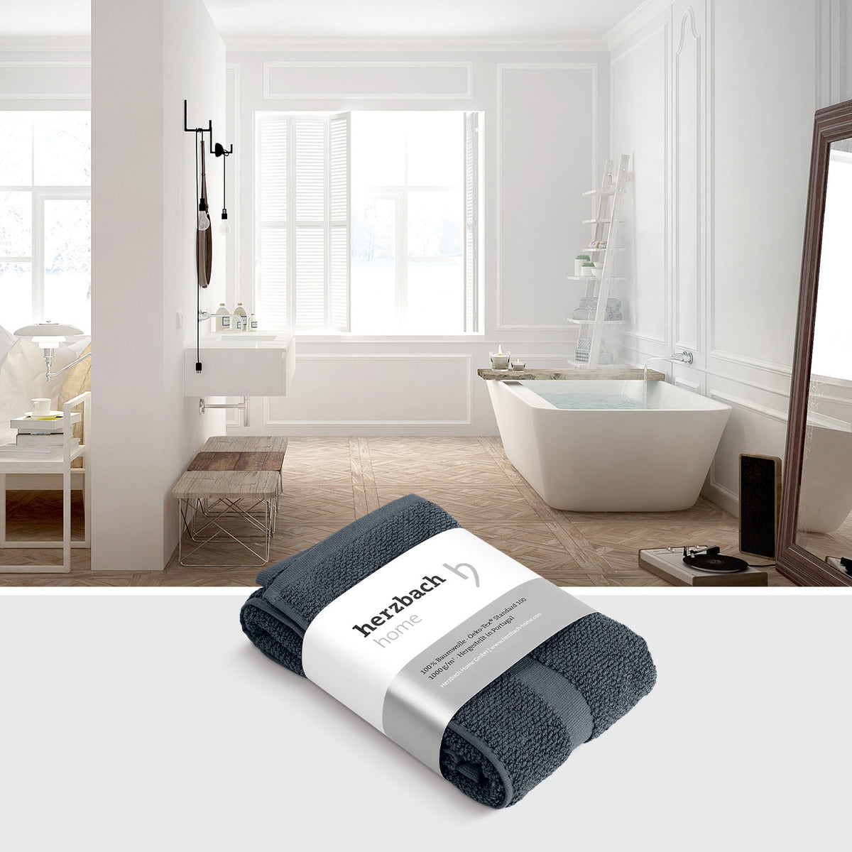 Spa Badvorleger Anthrazit – gefalteter Badvorleger mit herzbach home Banderole vor hellem Lifestyle-Badezimmer mit freistehender Badewanne und Holzparkett