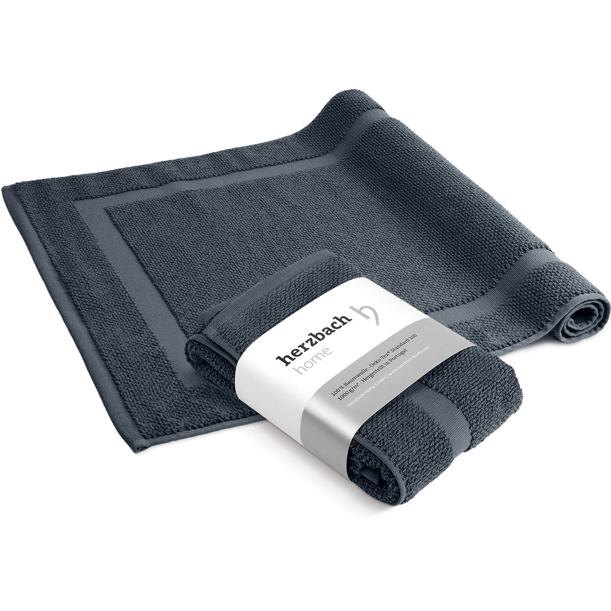 Spa Badvorleger Anthrazit 2er Set – ausgebreiteter und mit herzbach home Banderole aufgerollter Badvorleger von schräg oben, anthrazitfarbenes Baumwoll-Frottier mit Rahmendesign, weißer Hintergrund, 50 x 80 cm