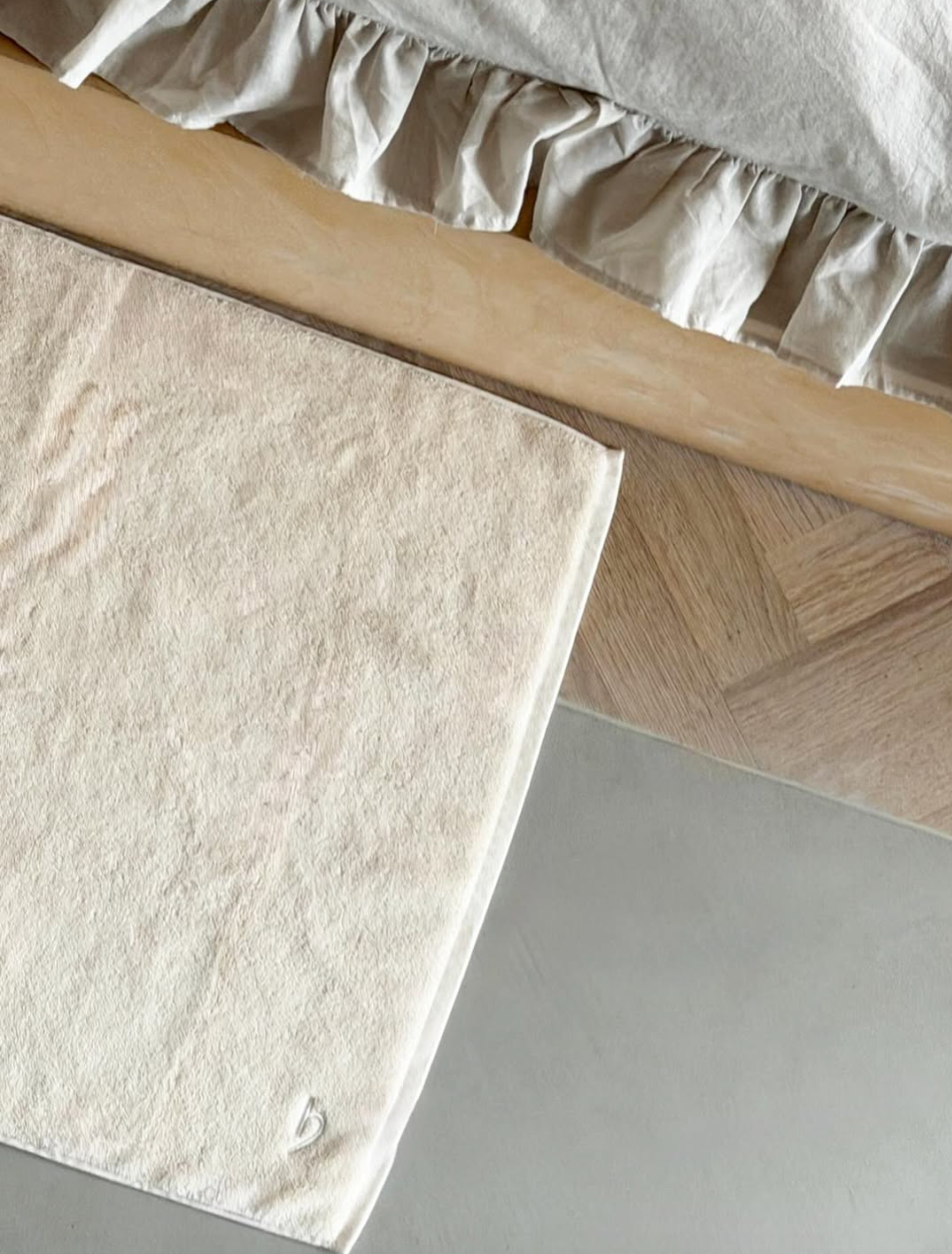 Herzbach home Badvorleger vor einem Bettgestell aus hellem Holz – weicher Frottier-Vorleger auf Holzboden mit Rüschenbettwäsche im Hintergrund