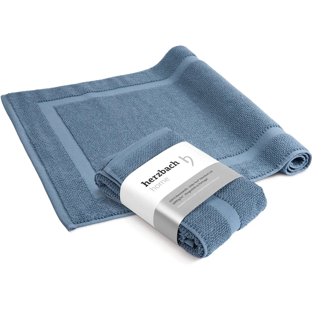 Spa Badvorleger Blau 2er Set – ausgebreiteter und mit herzbach home Banderole aufgerollter Badvorleger von schräg oben, stahlblaues Baumwoll-Frottier mit Rahmendesign, weißer Hintergrund, 50 x 80 cm