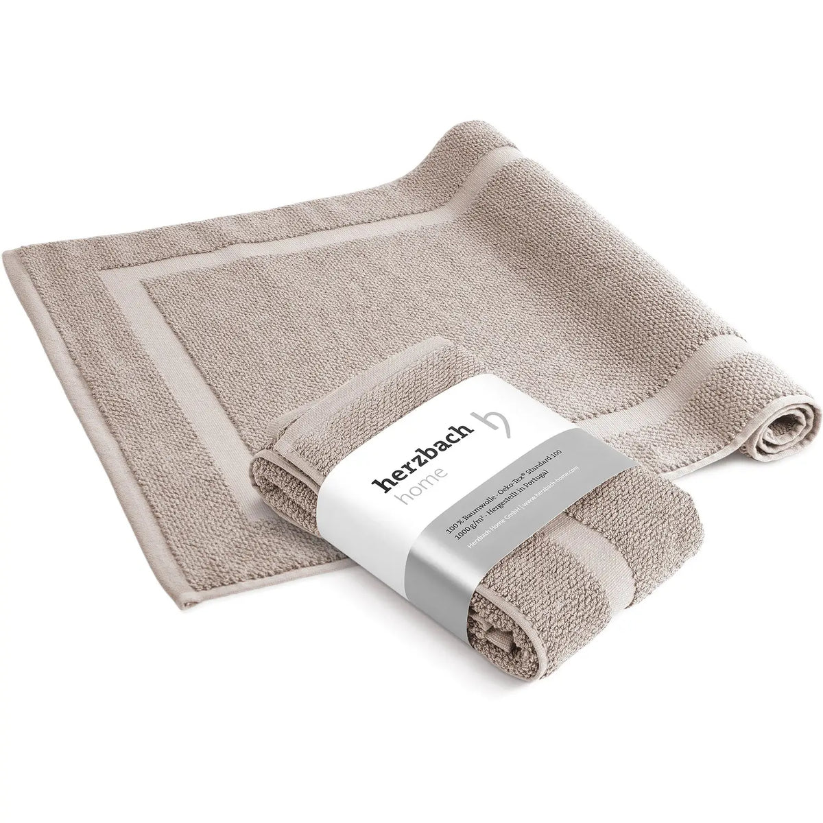 Spa Badvorleger Sandgrau 2er Set – ausgebreiteter und mit herzbach home Banderole aufgerollter Badvorleger von schräg oben, sandgraues Baumwoll-Frottier mit Rahmendesign, weißer Hintergrund, 50 x 80 cm