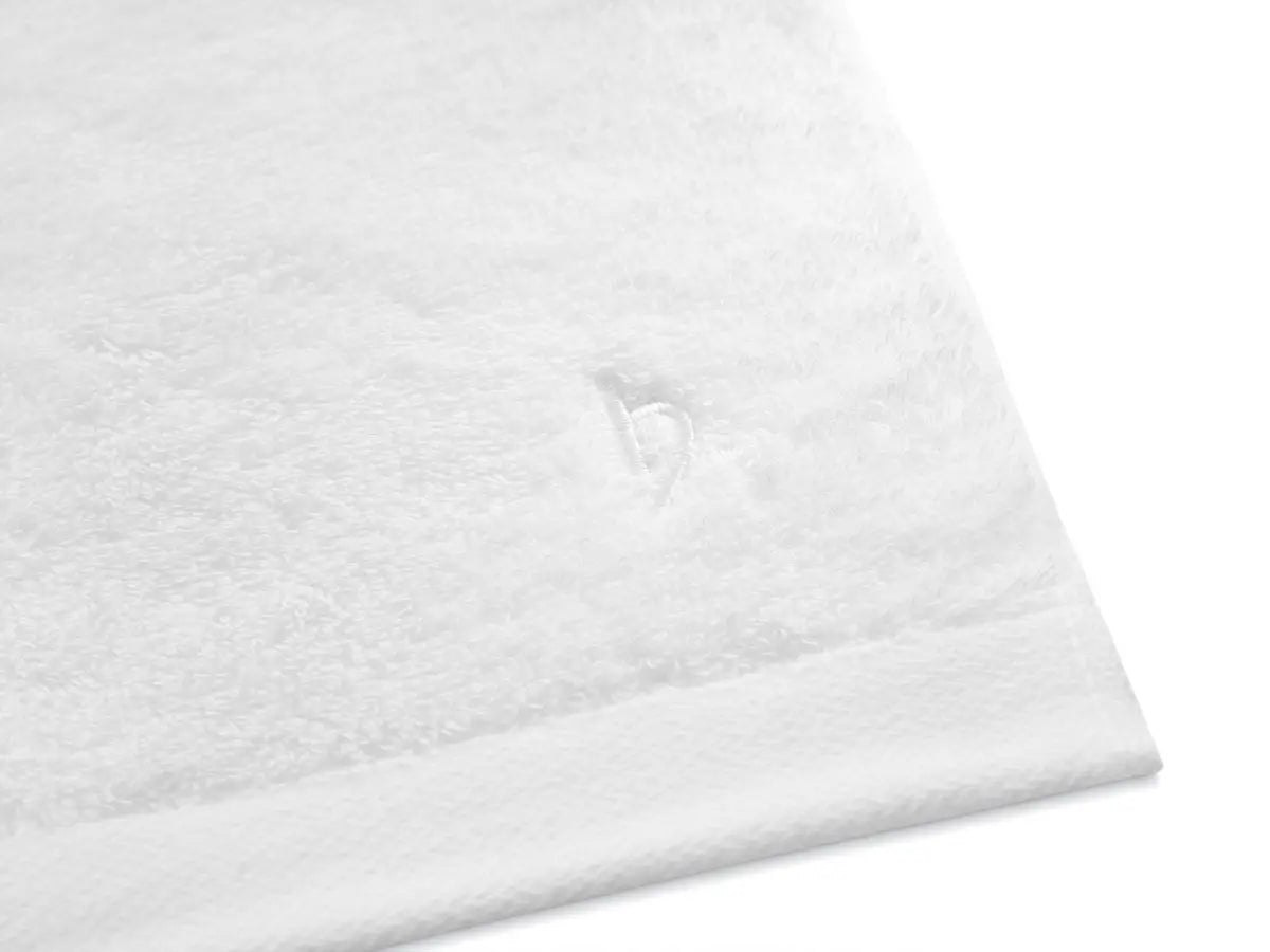 Eckdetail von weißem Pura Frottier mit eingesticktem Herz-Logo und glattem Saum
Schrägansicht