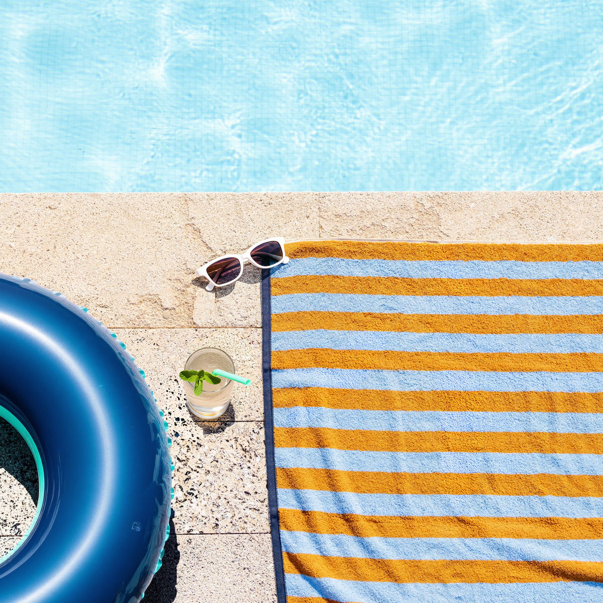 Badetuch Good Vibes Caramel Hellblau am Poolrand ausgebreitet – Sommerfeeling von oben mit Schwimmring, Sonnenbrille und Erfrischungsgetränk