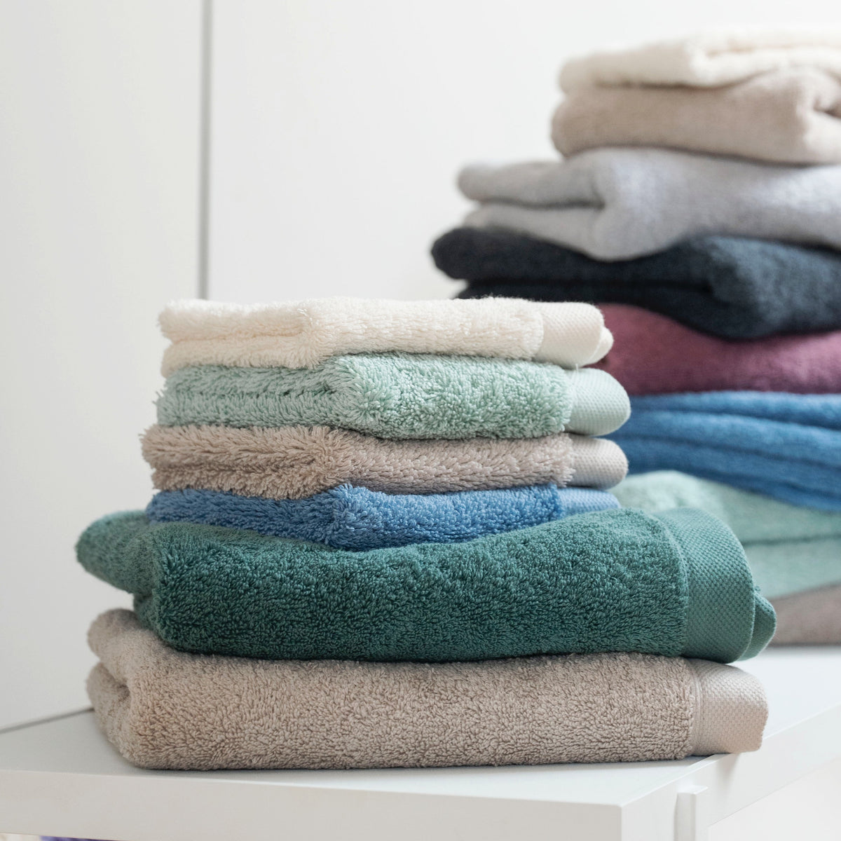 Pura Frottier-Handtücher in allen Farben – zwei Stapel auf weißer Ablage, im Vordergrund Ozeangrün und Sandgrau, im Hintergrund die gesamte Farbpalette von Creme bis Anthrazit