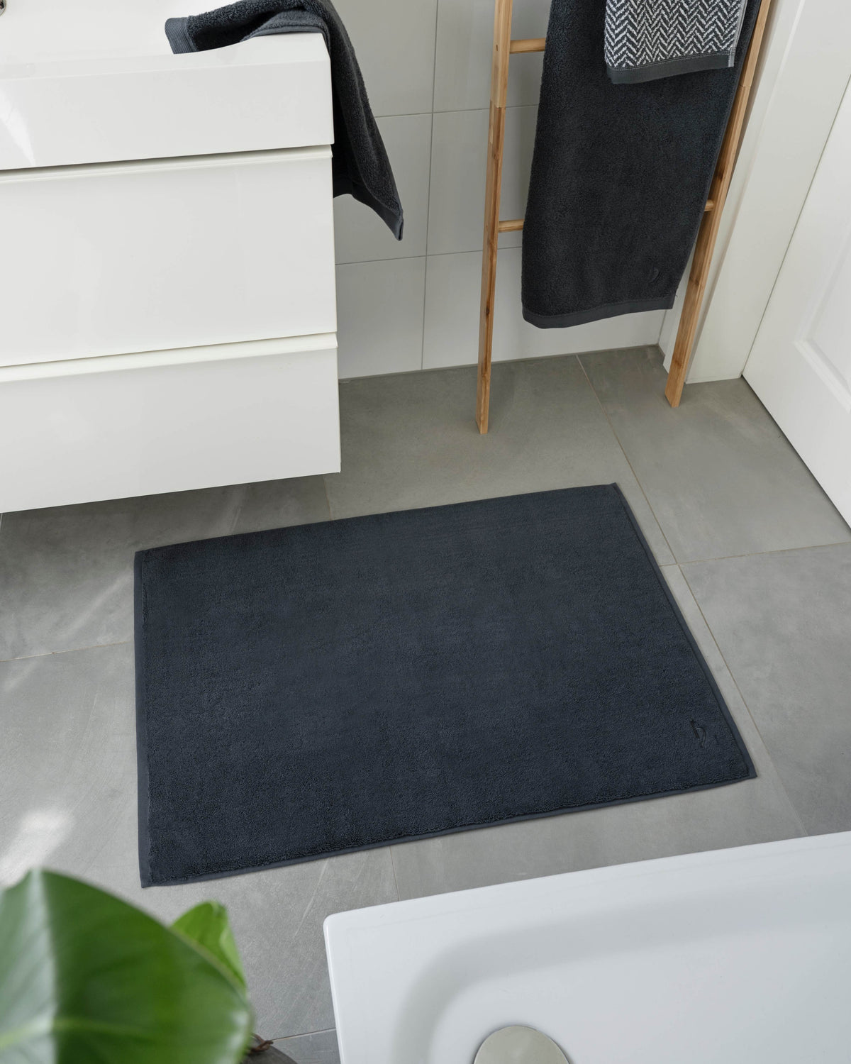 Pura Badvorleger Anthrazit – Lifestyle-Vogelperspektive im modernen weißen Badezimmer zwischen Waschtisch und Badewanne, kombiniert mit anthrazitfarbenen Frottier-Handtüchern auf Holzleiter, graue Bodenfliesen, grüne Zimmerpflanze