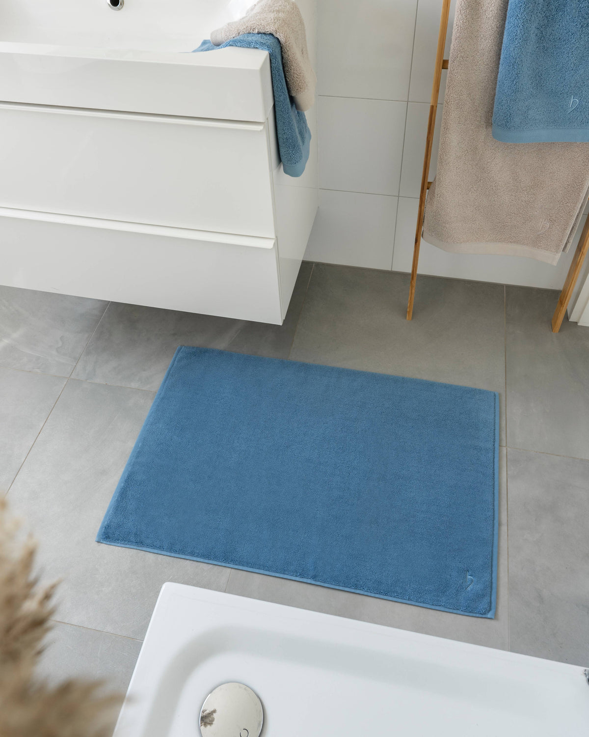 Pura Badvorleger Blau – Lifestyle-Vogelperspektive im modernen Badezimmer vor Waschtisch und Badewanne, kombiniert mit blauen Frottier-Handtüchern auf Goldleiter, graue Bodenfliesen, Pampasgras-Deko