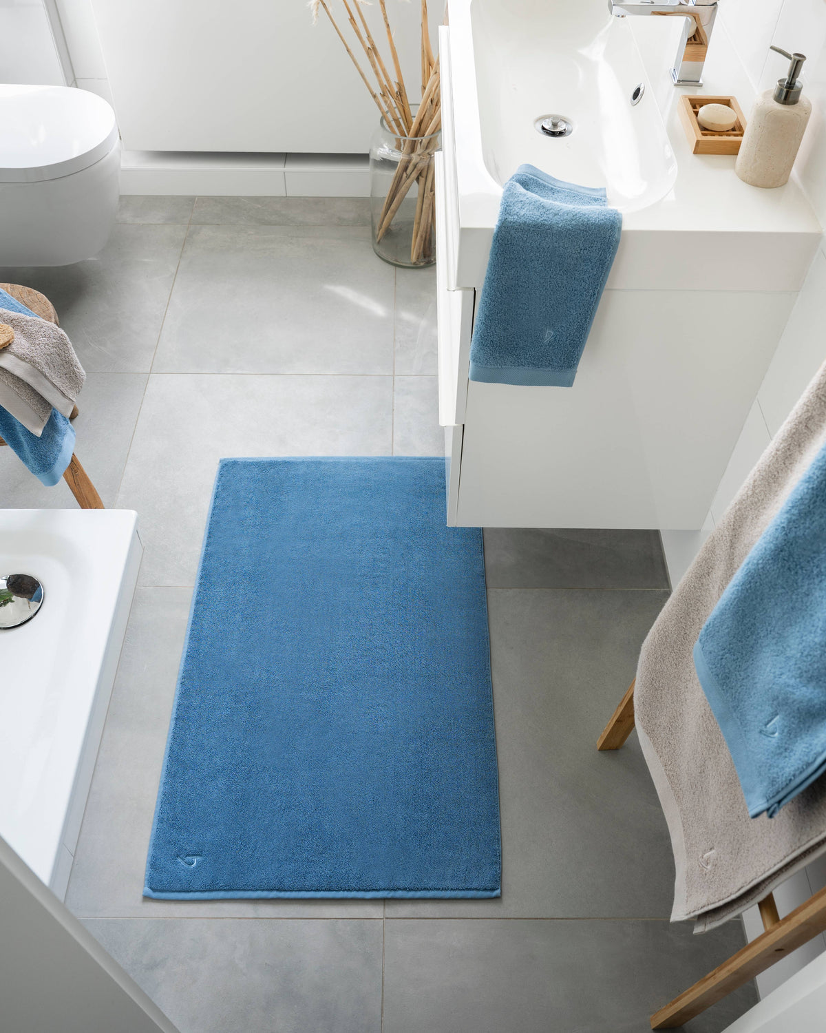 Pura Badvorleger Blau – Lifestyle-Draufsicht im hellen modernen Badezimmer zwischen Badewanne und Waschtisch, kombiniert mit blauen Frottier-Handtüchern auf Holzstuhl und Handtuchhalter, Bambus-Deko und Seifenspender