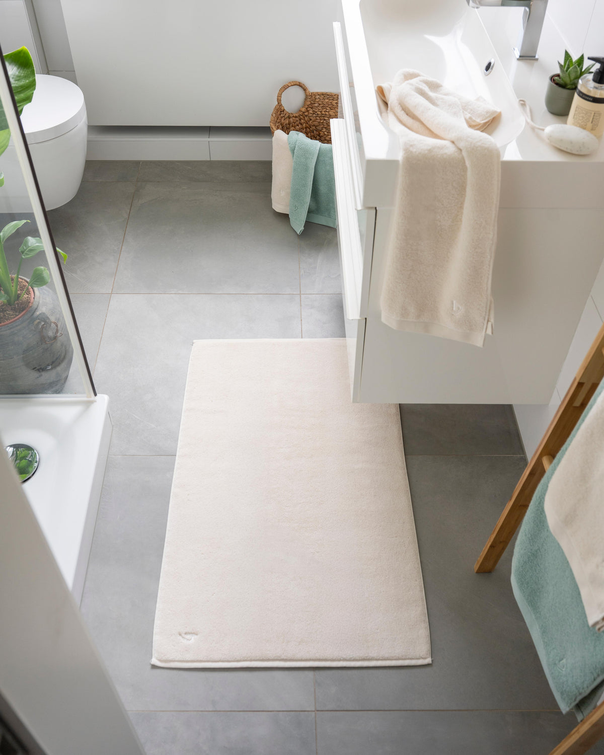 Pura Badvorleger Creme – Lifestyle-Draufsicht im modernen Badezimmer vor Dusche und Waschtisch, kombiniert mit cremefarbenen und mintgrünen Handtüchern auf Holzleiter