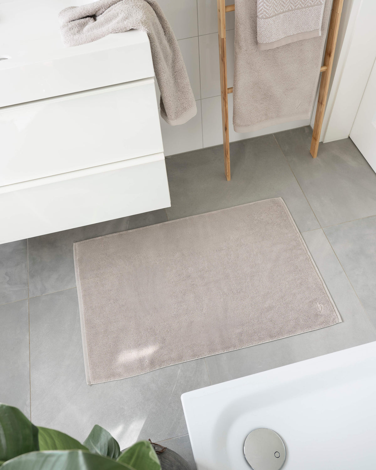 Pura Badvorleger Sandgrau – Lifestyle-Vogelperspektive im modernen Badezimmer zwischen Waschtisch und Badewanne, kombiniert mit sandgrauen Frottier-Handtüchern auf Holzleiter, graue Bodenfliesen