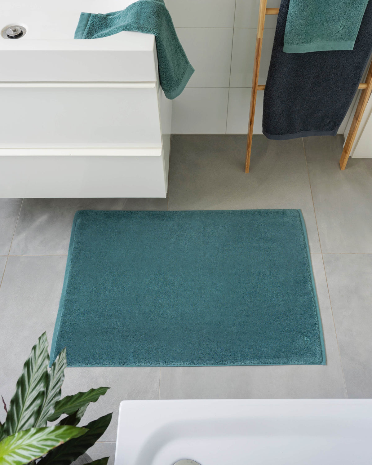 Pura Badvorleger Ozeangrün – Lifestyle-Vogelperspektive im modernen Badezimmer zwischen Waschtisch und Badewanne, kombiniert mit ozeangrünen und anthrazitfarbenen Frottier-Handtüchern auf Holzleiter, graue Bodenfliesen, grüne Zimmerpflanze