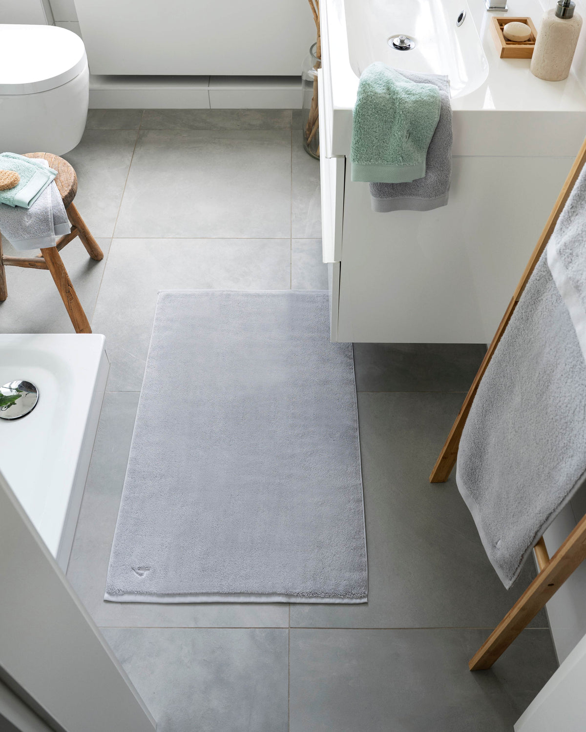 Pura Badvorleger Silbergrau – Lifestyle-Ansicht von oben im hellen Badezimmer zwischen Waschtisch und Badewanne, kombiniert mit mintgrünen und silbergrauen Frottier-Handtüchern auf Holzleiter