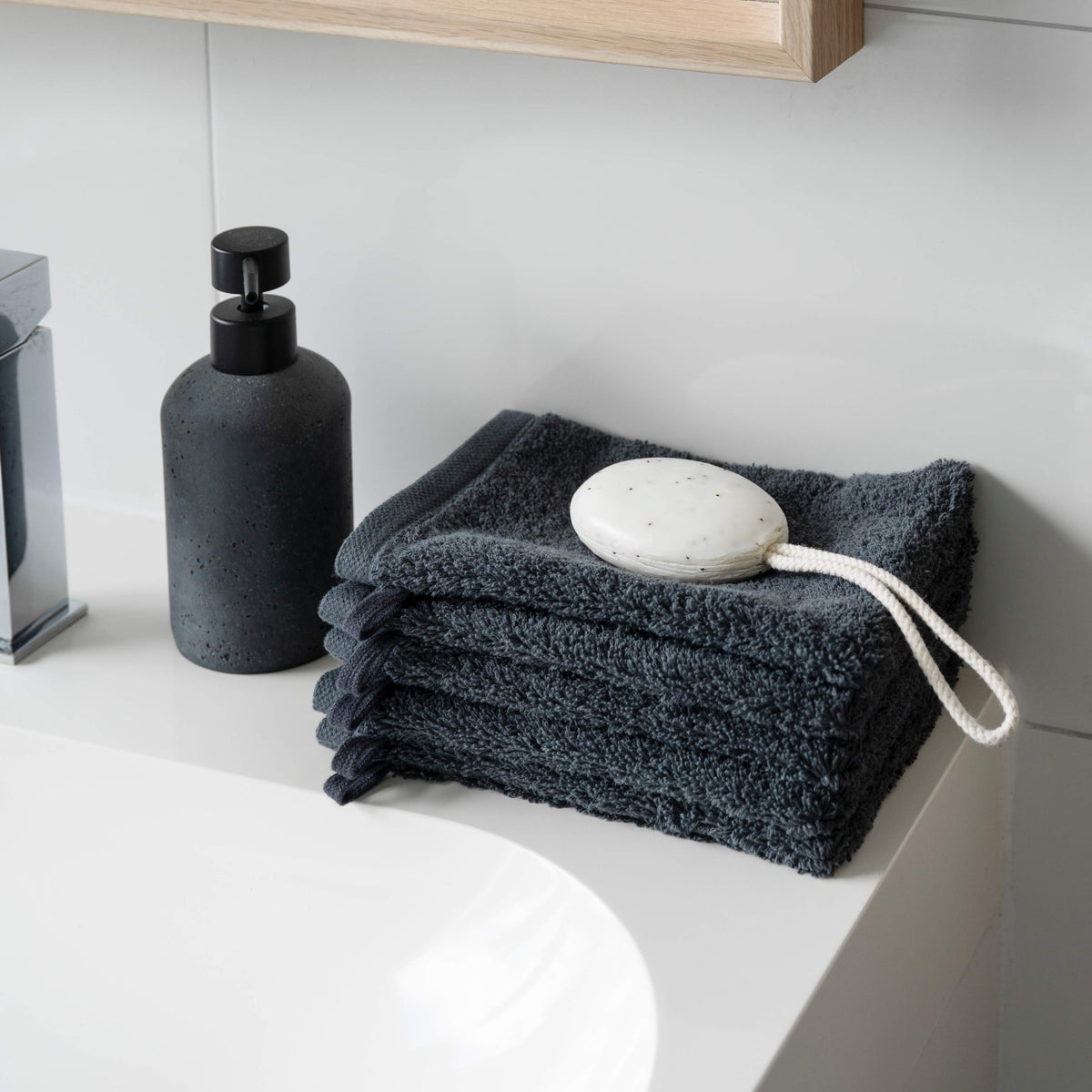 Waschhandschuhe Pura Anthrazit gestapelt auf dem Waschtisch – neben schwarzem Keramik-Seifenspender, Naturseife mit Baumwollschnur obenauf, modernes Badezimmer-Ambiente