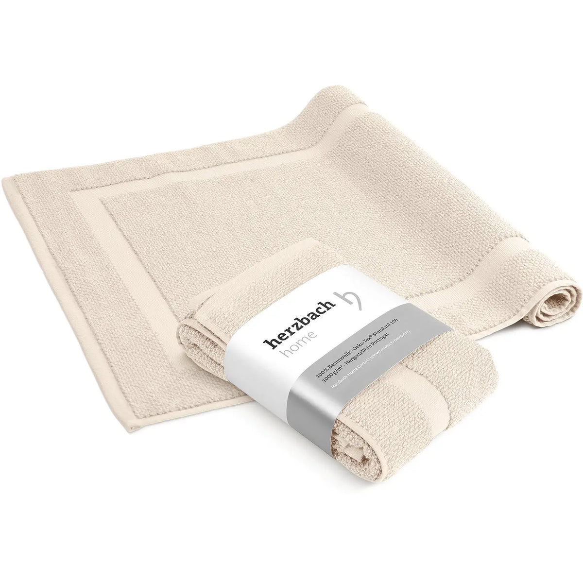 Spa Badvorleger Creme 2er Set – ausgebreiteter und mit herzbach home Banderole aufgerollter Badvorleger von schräg oben, cremefarbenes Baumwoll-Frottier mit Rahmendesign, weißer Hintergrund, 50 x 80 cm