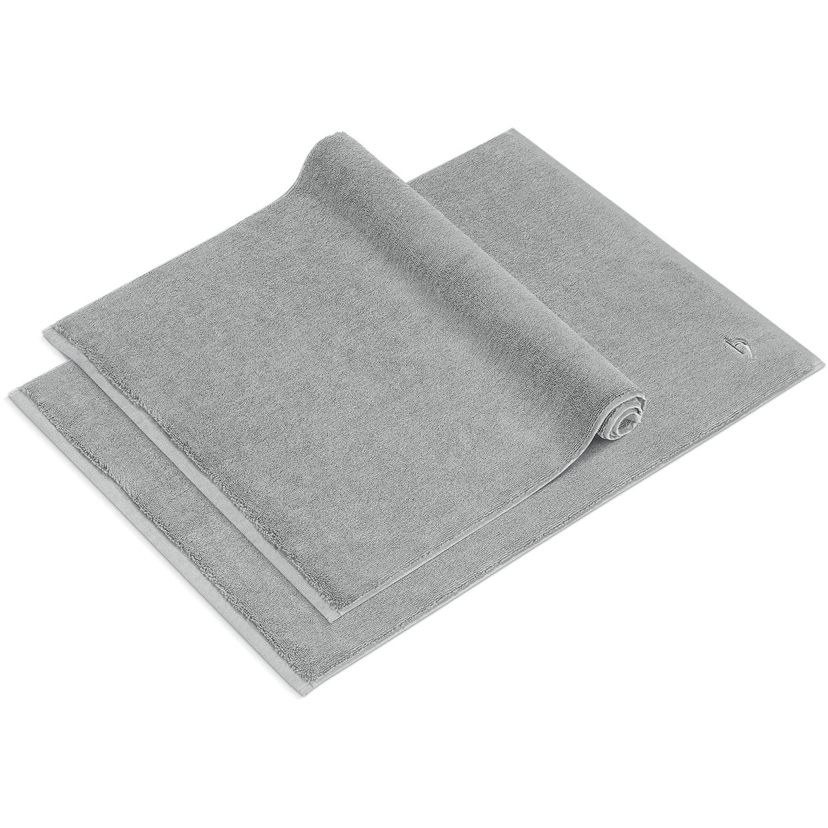 Pura Badvorleger 2er Set Silbergrau – ein ausgebreiteter und ein halb aufgerollter Badvorleger 50x70 cm von schräg oben, dichtes Frottier mit feingemusterter Bordüre und Ton-in-Ton-Logostickerei, 100% Baumwolle 1200 g/m²