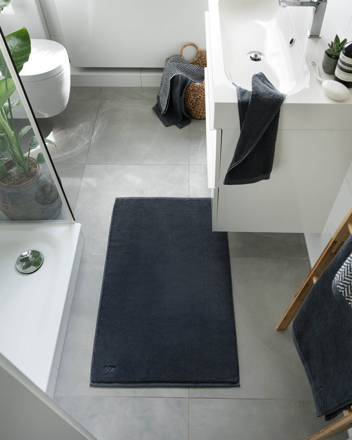 Pura Badvorleger Anthrazit – Lifestyle-Vogelperspektive im hellen Badezimmer zwischen Dusche und Waschtisch, kombiniert mit anthrazitfarbenen Frottier-Handtüchern in Weidenkorb und auf Holzregal, Zimmerpflanzen und Deko-Accessoires