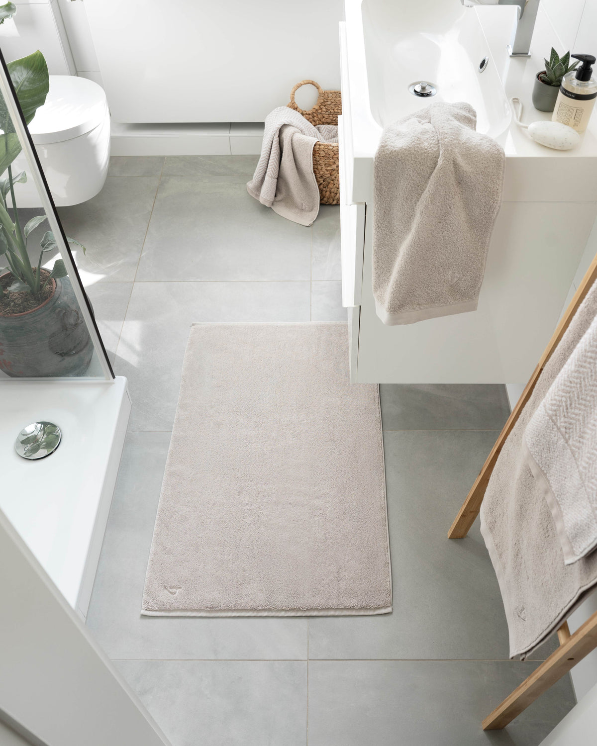 Pura Badvorleger Sandgrau – Lifestyle-Draufsicht im hellen weißen Badezimmer vor Dusche und Waschtisch, kombiniert mit sandgrauen Handtüchern in Weidenkorb und auf Holzleiter, grüne Zimmerpflanze
