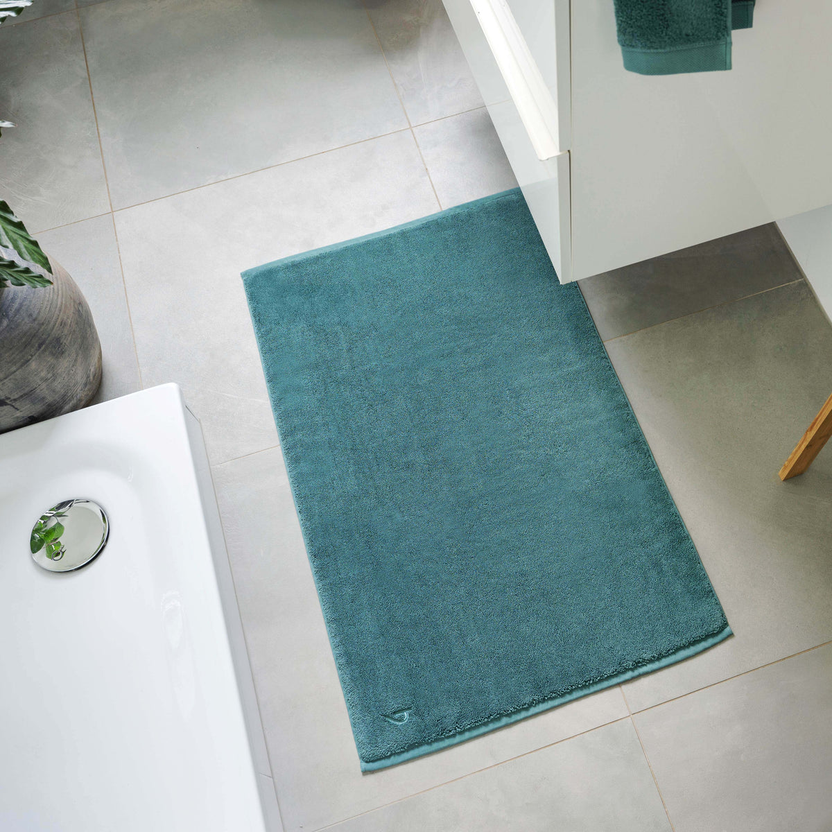 Pura Badvorleger Ozeangrün – Lifestyle-Draufsicht im hellen Badezimmer neben freistehender Badewanne und Waschtisch, kombiniert mit ozeangrünen Frottier-Handtüchern auf Holzleiter, große grüne Zimmerpflanze und Deko-Vase