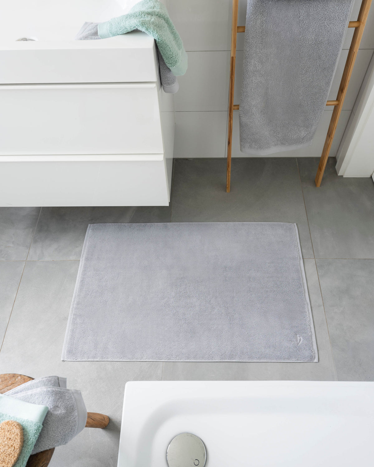 Pura Badvorleger Silbergrau – Lifestyle-Draufsicht im modernen weißen Badezimmer vor freistehender Badewanne, kombiniert mit mintgrünen Handtüchern auf Holzleiter, grauer Fliesenboden
