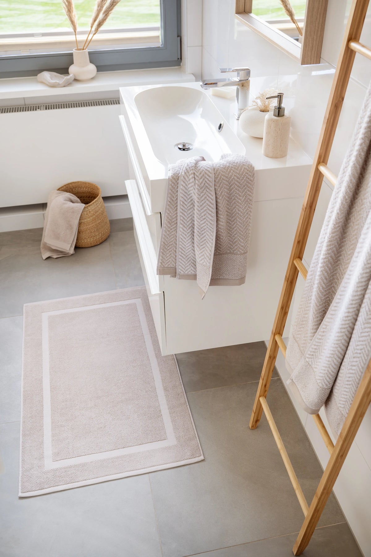 Spa Badvorleger Sandgrau – Lifestyle-Schrägansicht im hellen modernen Badezimmer vor Waschtisch, ausgebreiteter Badvorleger mit Rahmendesign, Bambusleiter mit sandgrauen Handtüchern, Weidenkorb und Deko-Vase am Fenster